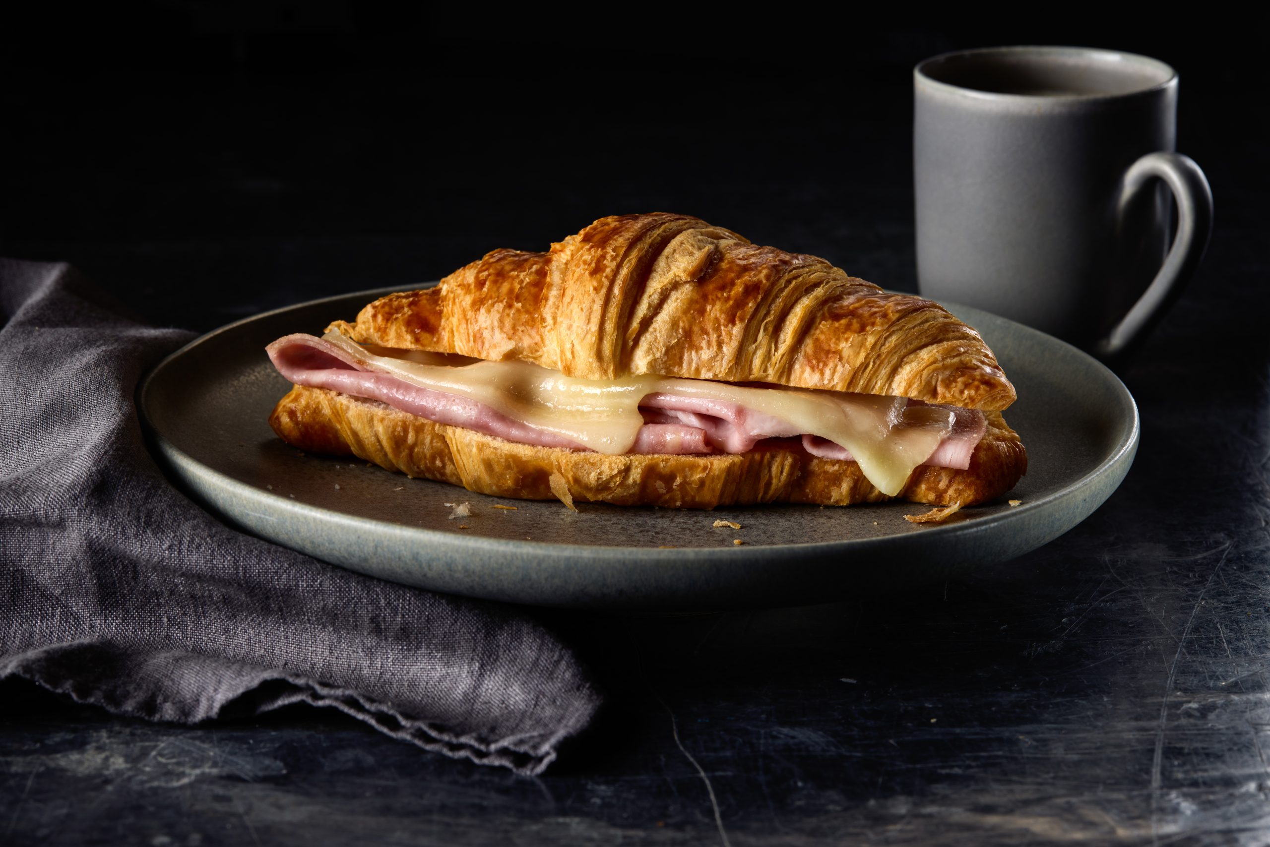 Golden ham and cheese croissant with coffee on grey plate, cosy minimalist breakfast setting.
